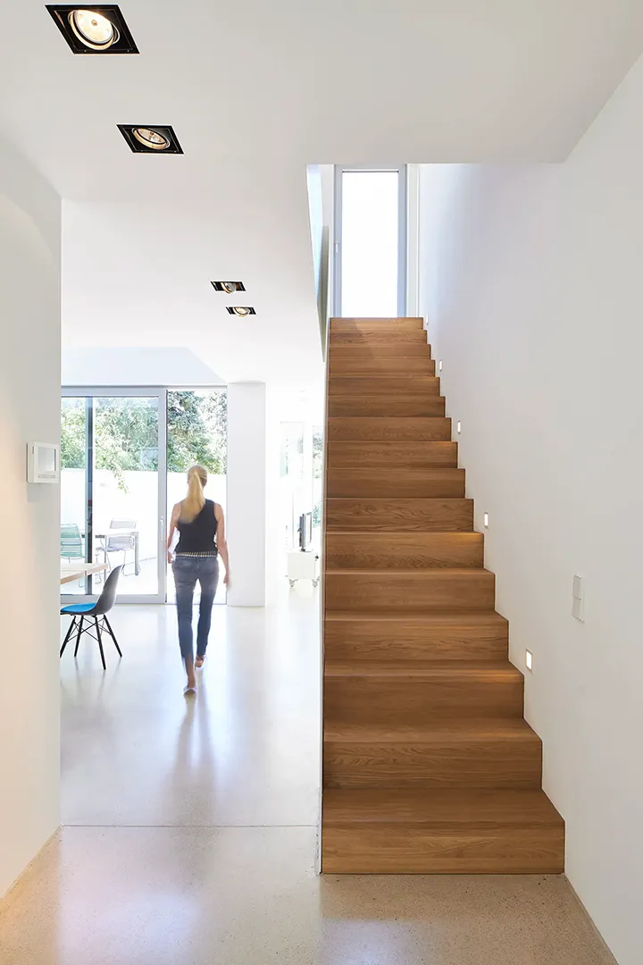 Einläufige Holztreppe mit filigraner weißer Stahlbrüstung, das warme Holz setzt sich gegen den weißen Putz und den hellen geschliffenen Estrichboden ab.