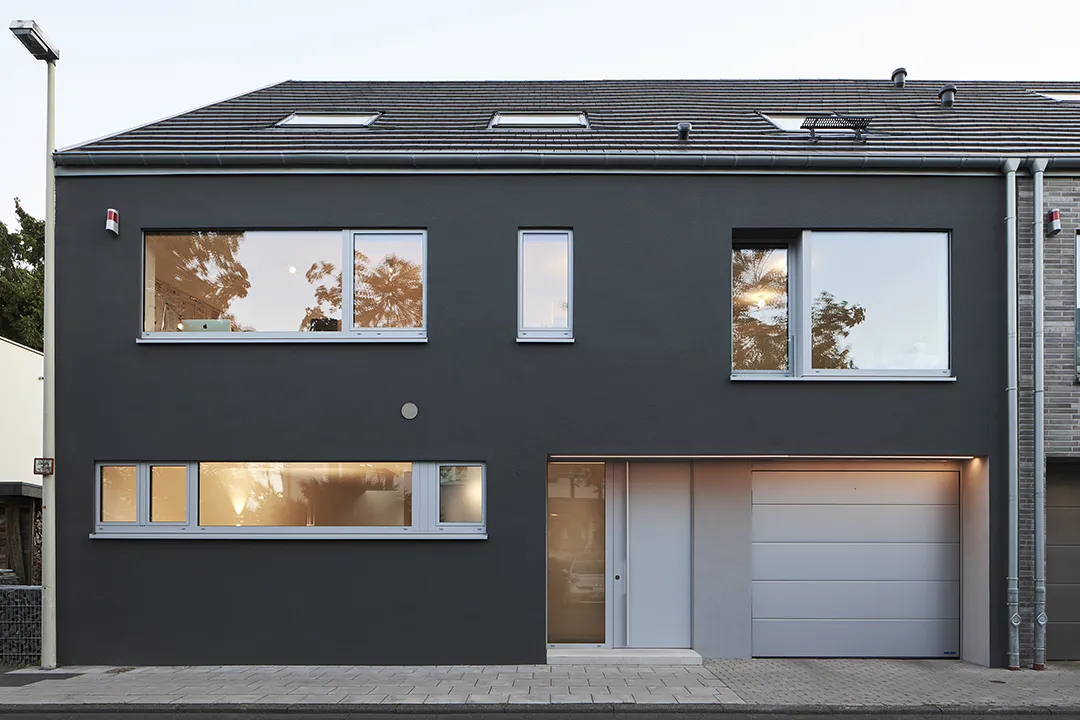 Straßenseitige Außenansicht eines Reihenhauses mit dunkel verputzter Lochfassade und asymmetrisch gesetzten Rahmenfenstern; Eingangsbereich und Garagentor sind flächenbündig in die Putzfläche eingeschnitten.