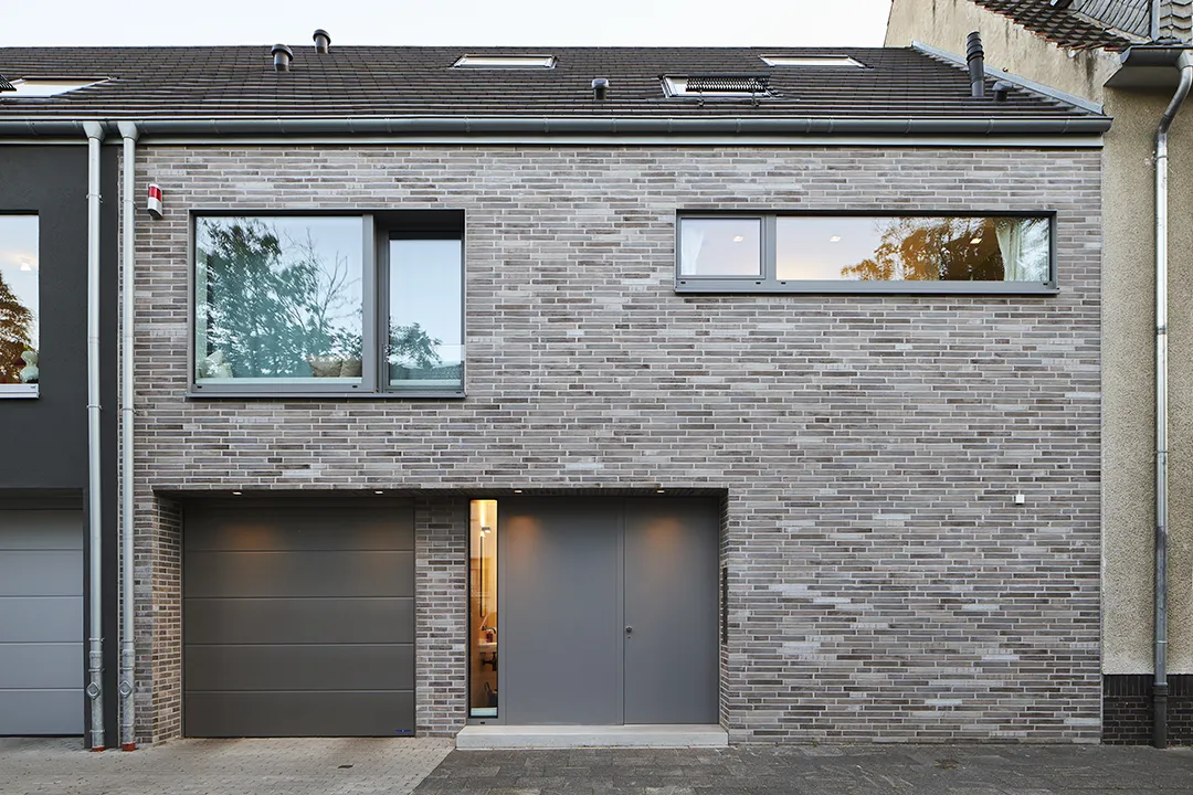 Straßenseitige Außenansicht eines Reihenhauses mit durchgehender Riemchenfassade, Garagentore und Haustür sind in dunklem Metall flächenbündig eingelassen, die Lochfassade setzt querformatige Fenster in gleichmäßigem Rhythmus.
