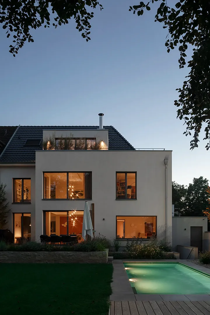 Gartenseitige Ansicht zur blauen Stunde, kubischer Putzbaukörper mit Lochfassade vor dem geneigten Bestandsdach, zurückgesetzte Dachterrasse mit Pflanztrögen, im Vordergrund Schwimmbecken und Natursteinstützmauer.