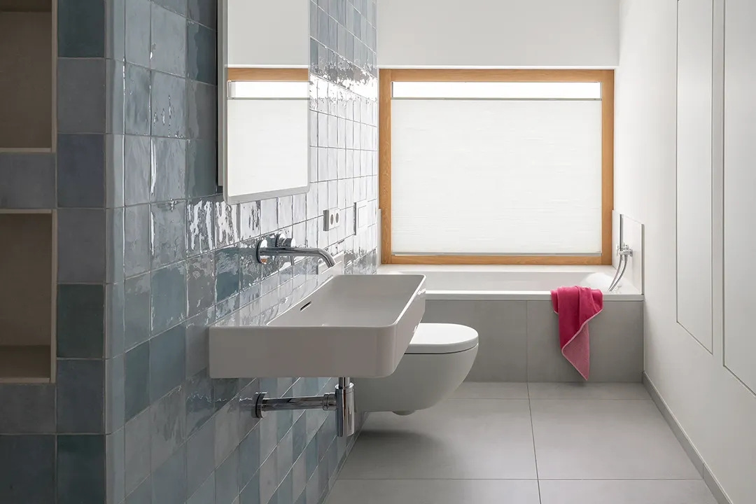 Bad mit handgeformtem Fliesenspiegel an der Installationswand, wandhängender Waschtisch und WC, raumbreit eingebaute Wanne unter dem Fenster mit Holzlaibung und Plissee.