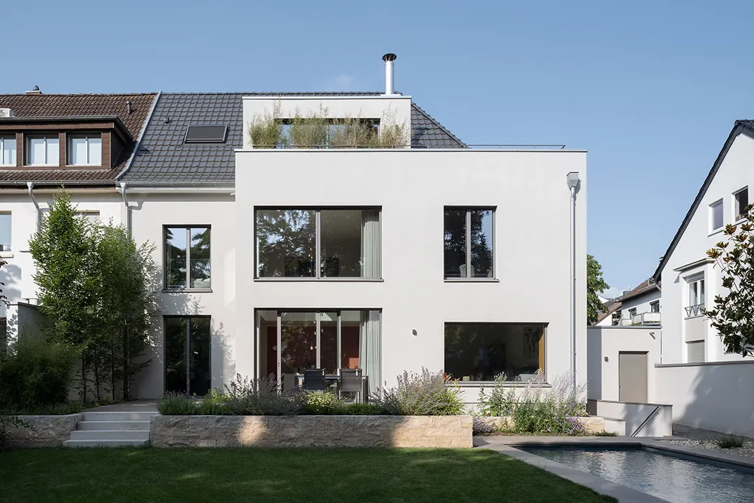 Gartenseitige Gesamtansicht des Anbaus als kubische Aufstockung vor dem Bestandsdach, differenzierte Fensterformate über zwei Geschosse, Terrassierung mit Natursteinmauer und Schwimmbecken.