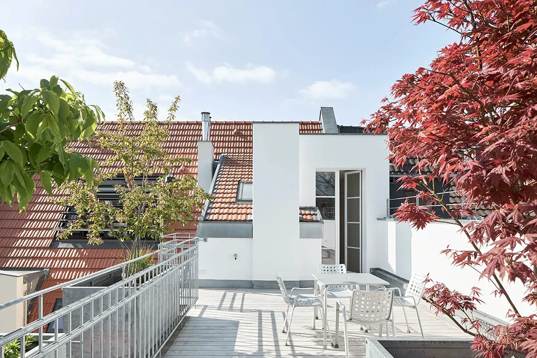 Dachterrasse mit Holzbelag und Metallgeländer zwischen weißen Wandscheiben, ringsum Ziegeldächer der Nachbarhäuser und ein roter Ahorn.