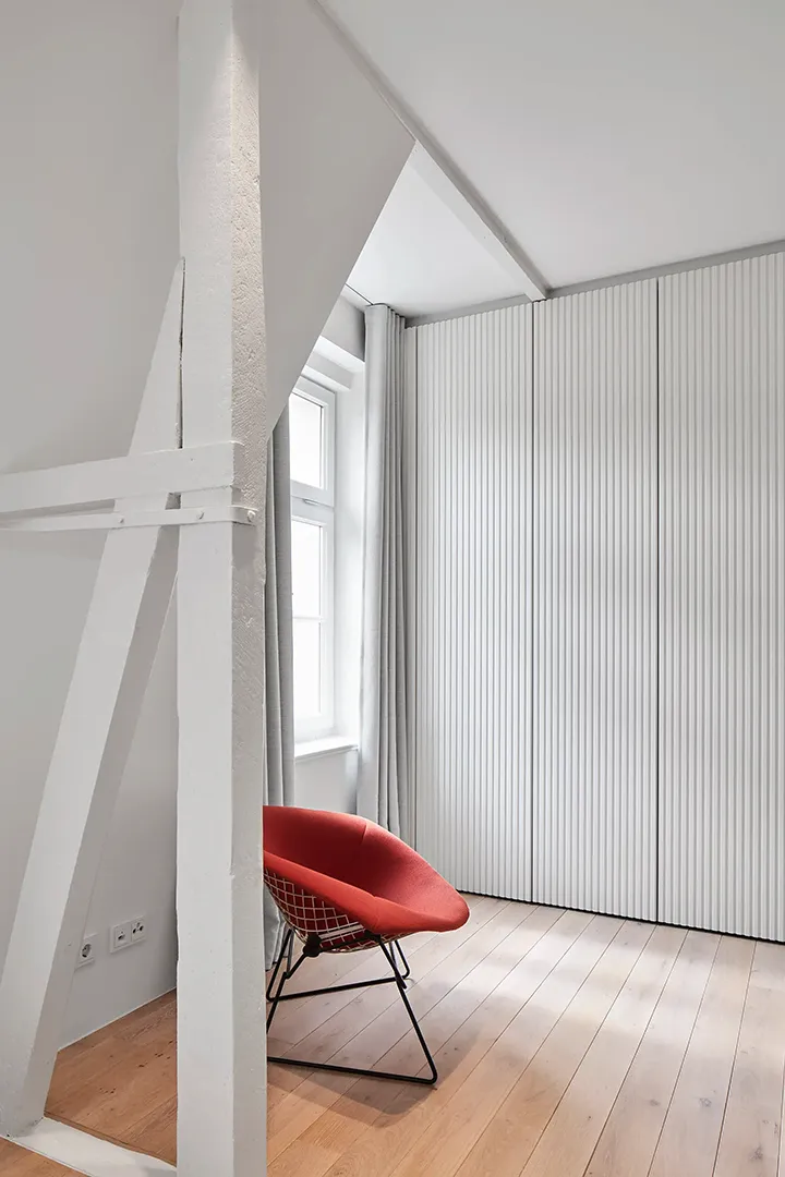Schlafzimmer mit weiß gestrichenem Fachwerkständer im Vordergrund, dahinter eine geriffelte Schrankfront und ein Fenster mit hellem Vorhang.