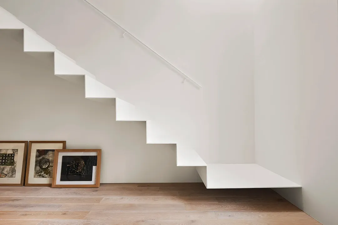 Freitragende weiße Faltwerktreppe mit schlankem Rundhandlauf an der Wand, darunter mehrere gerahmte Grafiken auf dem Holzboden.