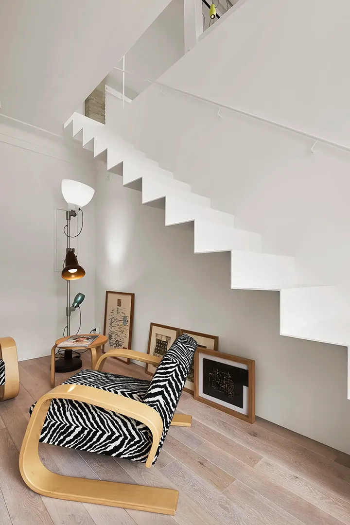 Wohnbereich unter der freitragenden weißen Treppe, davor ein Sessel mit gemustertem Bezug und angelehnte Bilderrahmen.