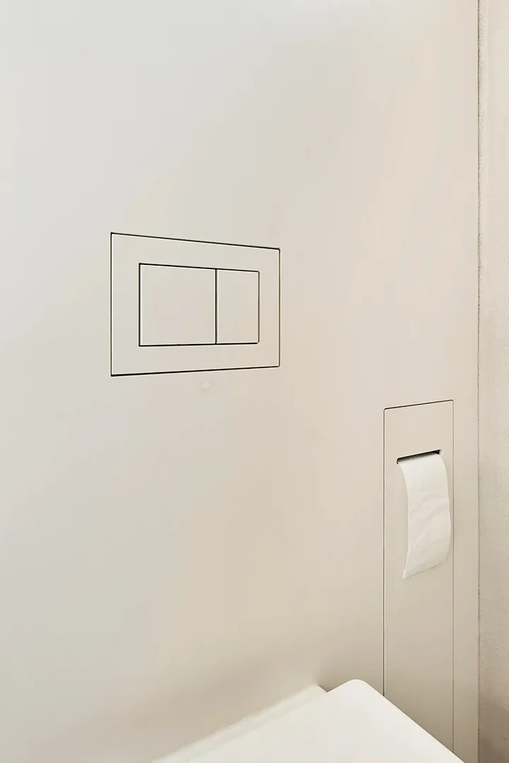 Detail einer fugenlos gefliesten Wand mit flächenbündig eingelassener Betätigungsplatte und in die Wand eingelassenem Papierhalter.