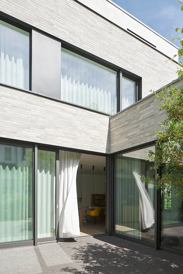 Über Eck geführte Verglasung im Erdgeschoss unter auskragendem Obergeschoss mit Klinkerriemchen-Verkleidung, geöffnetes Schiebeelement mit im Wind stehendem Vorhang, gepflasterter Terrassenbelag.