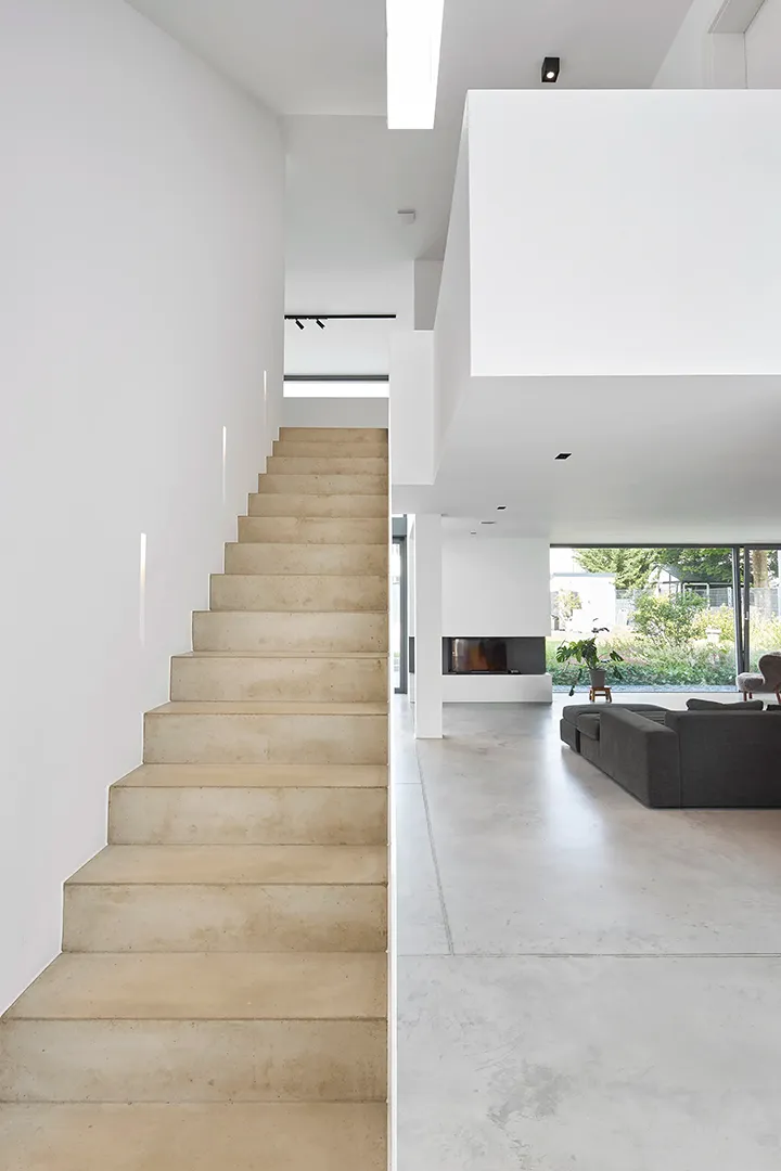 Einläufige Sichtbetontreppe an geschlossener Stahlbrüstung mit eingelassenen Lichtvouten, rechts der offene Wohnbereich mit Kaminwand und geschliffenem Estrich.