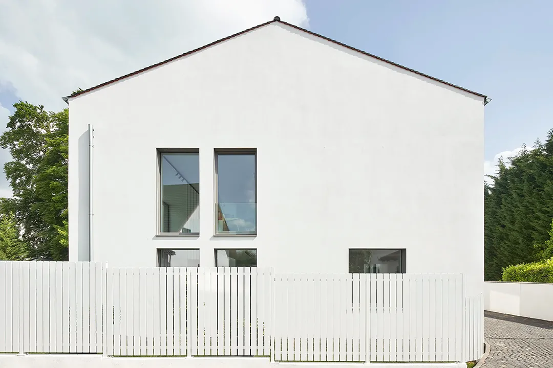 Frontale Giebelansicht als geschlossene Putzfläche mit gestaffelten Fensteröffnungen, davor vertikal gegliederte Holzeinfriedung.
