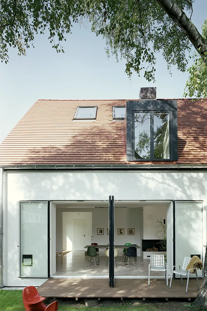 Gartenseitige Außenansicht des eingeschossigen Hauses mit Satteldach; eine raumhohe Faltglasfassade mit dunklem Metallrahmen öffnet den Wohnbereich vollständig zur Holzterrasse, im Dach sitzt ein dunkler Gaubenaufbau mit Rahmenverglasung.