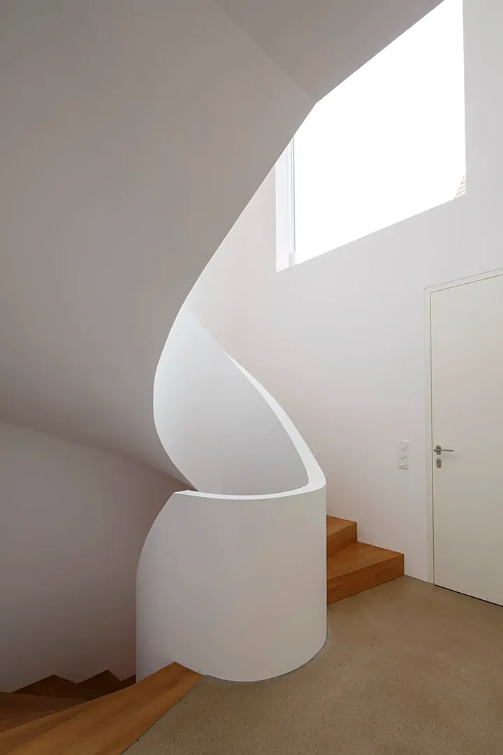 Freigeformte Wendeltreppe aus weißem Putz mit Holzstufen windet sich durch einen schmalen, oben belichteten Schacht; die plastisch modellierte Brüstungswange gibt dem Treppenraum eine skulpturale Qualität.