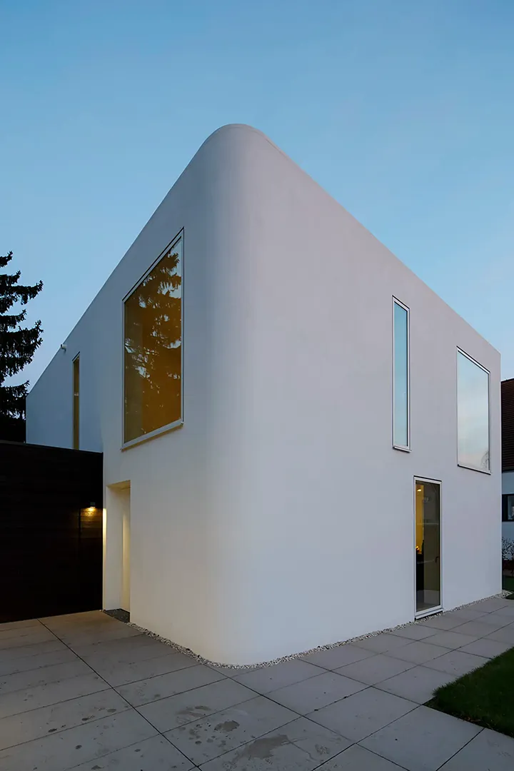 Seitenansicht des Baukörpers in der Abenddämmerung; die gerundete Gebäudeecke verbindet Eingangs- und Gartenseite nahtlos, ein großformatiges Fenster im Obergeschoss und ein schlankes Hochformatfenster im Erdgeschoss setzen Akzente in der sonst geschlossenen Putzfläche.