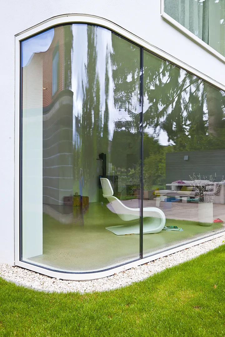 Bodentiefe Glasecke im Erdgeschoss mit gerundeter Eckverglasung ohne sichtbaren Pfosten; die geschwungene Oberkante der Öffnung folgt der Geometrie der Putzfassade und gibt den Blick in den Wohnraum frei.