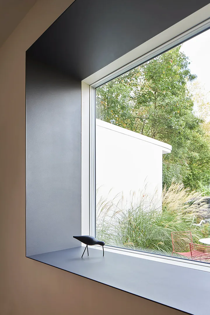 Tiefe, dunkel gefasste Laibung eines großformatigen Fensters mit Blick auf den begrünten Außenraum; auf der Fensterbank steht eine kleine Vogelfigur.