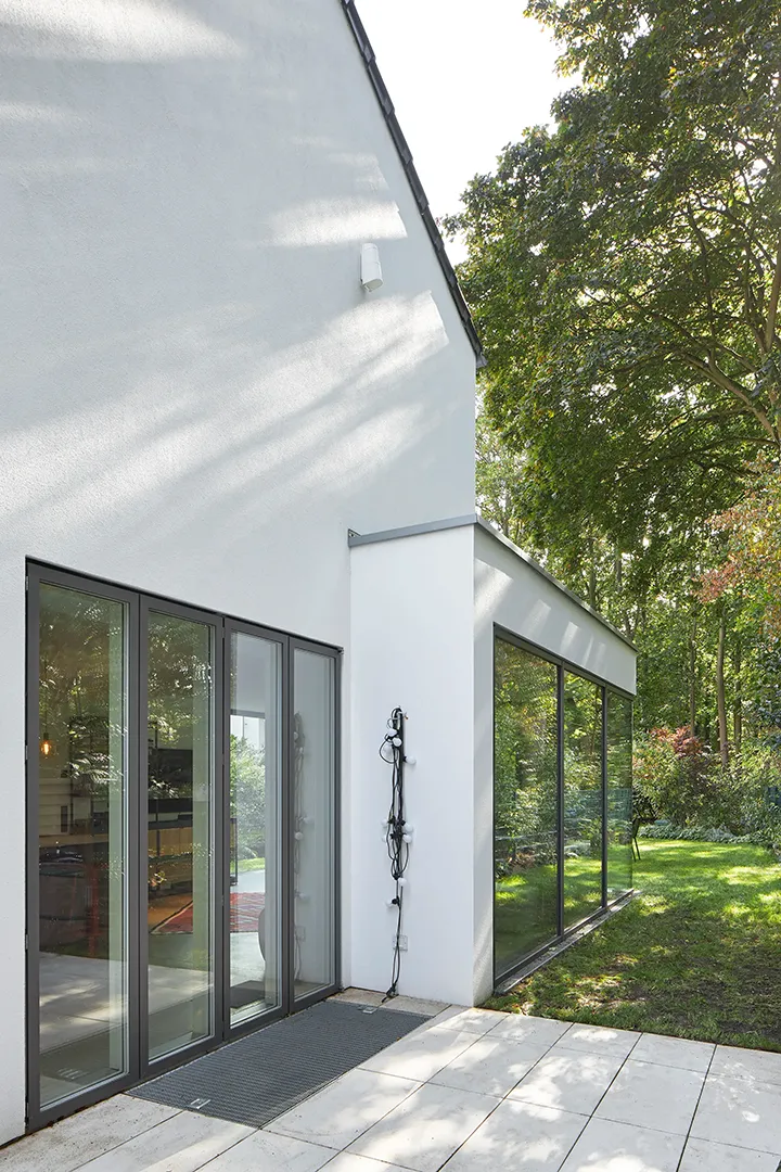 Gartenseitiges Fassadendetail mit Schiebeanlage aus dunklen Metallprofilen und anschließender bodentiefer Rahmenverglasung; die weiße Putzwandscheibe vermittelt zwischen den Öffnungen.