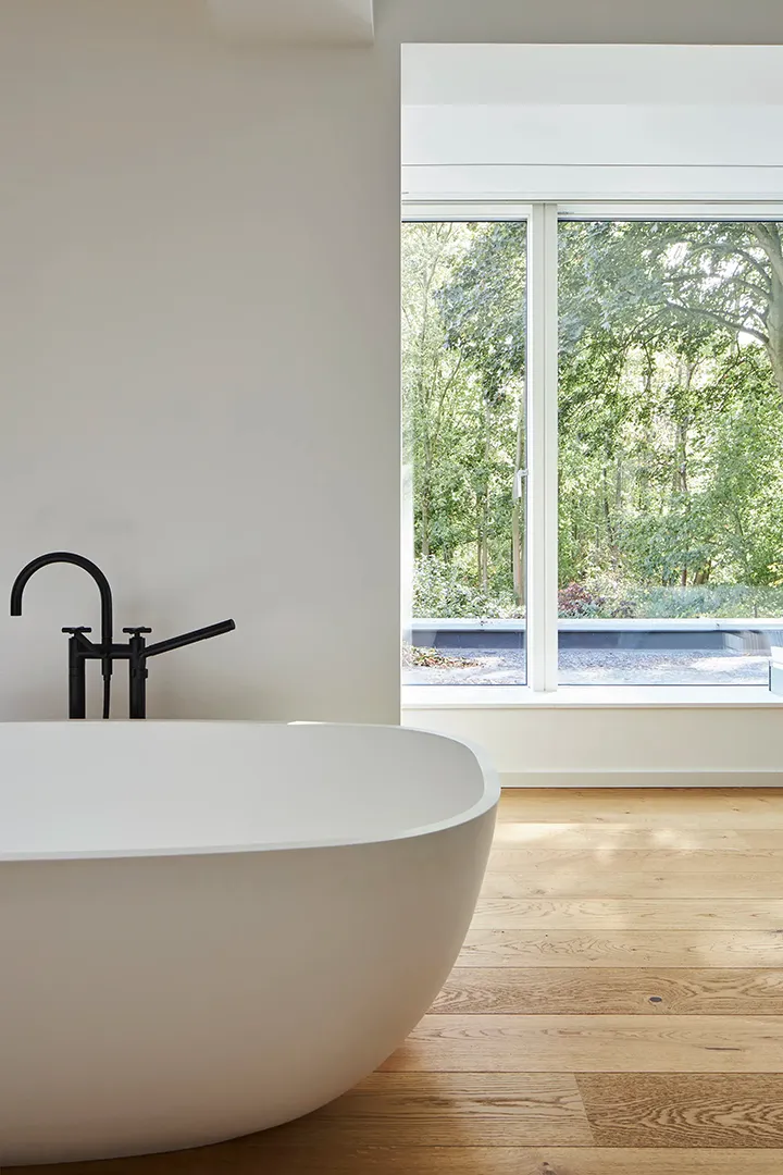 Freistehende Badewanne aus weißem Mineralwerkstoff auf Holzboden, mit schwarzer Standbatterie; das großzügige Fenster dahinter gibt den Blick in den bewachsenen Garten frei.