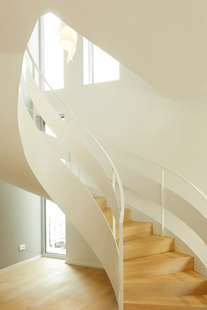 Freigeformte Wendeltreppe mit weißer Putzwange und Holzstufen windet sich durch einen hohen, oben belichteten Treppenraum; das einseitig geführte Metallgeländer folgt der Schwingung der Wandscheibe.
