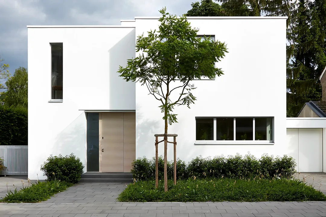 Straßenseitige Außenansicht eines zweigeschossigen Wohnhauses mit weißer Putzfassade und gestaffelten Baukörpern; eine flächenbündige Holztür sitzt in einer schmalen Einschnittlaibung, ein Hochformatfenster und ein liegendes Querformatfenster gliedern die geschlossene Wandfläche asymmetrisch.