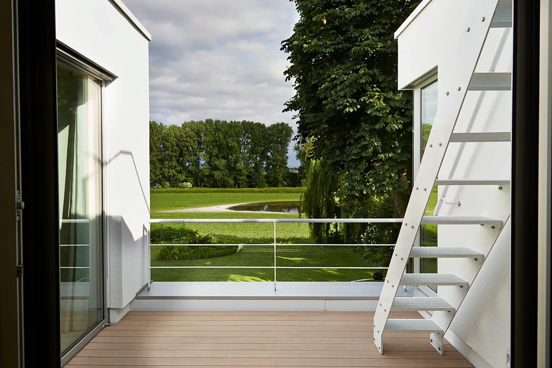Dachterrasse mit Holzbelag und schlichtem Metallgeländer zwischen zwei weißen Putzwandscheiben; eine außenliegende Metalltreppe führt auf das nächste Geschoss, der Blick öffnet sich auf Garten und Landschaft mit Teich.
