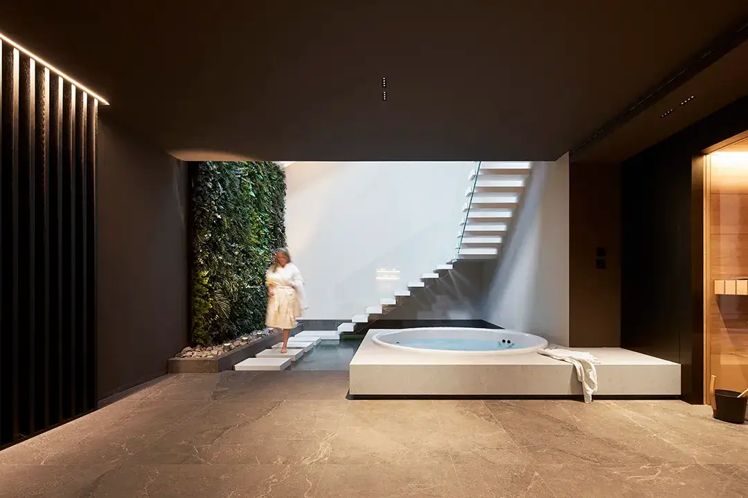 Blick durch den dunkel gefassten Wellnessbereich in den tageslichtbelichteten zweigeschossigen Raum mit Whirlpool und Wasserbecken, Pflanzenwand, auskragenden Sichtbetonstufen und Natursteinoberflächen.