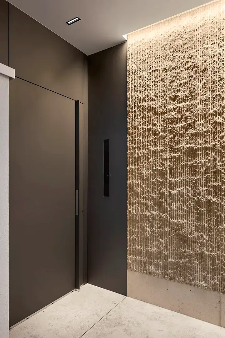 Eingangssituation mit flächenbündigem Türblatt in dunkler Wandverkleidung, integriertem Bedienfeld und angestrahlter Natursteinwand mit reliefartiger Oberfläche.