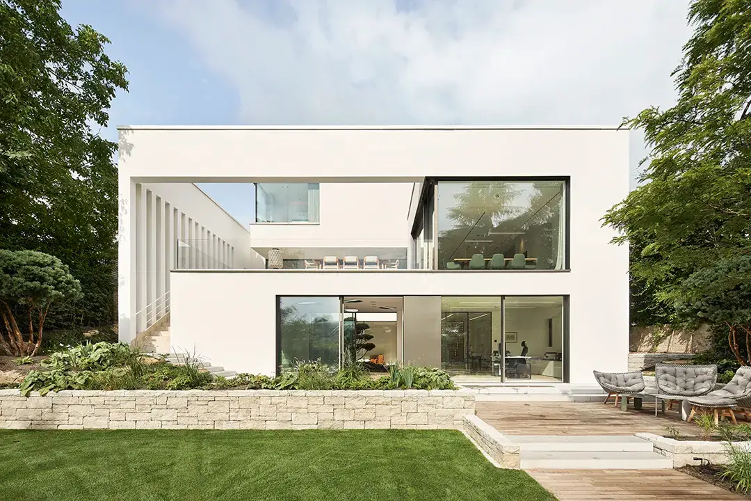 Gartenseitige Gesamtansicht mit tief eingeschnittener Terrasse im Erdgeschoss, durchlaufender Ganzglasbrüstung und geschosshoher Verglasung im Gartengeschoss, terrassiertes Gelände mit Natursteinstützmauer und Holzdeck.