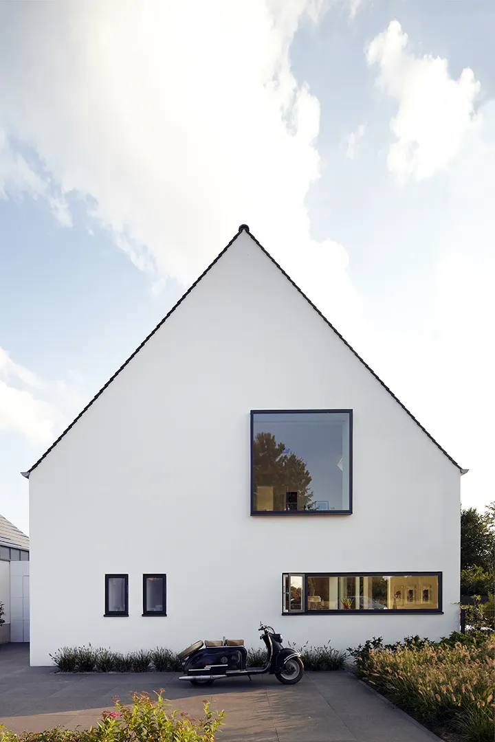 Traufseitige Außenansicht des Wohnhauses mit steilem Satteldach und geschlossener weißer Putzgiebelwand; ein einzelnes querformatiges Fenster und zwei schmale Lochöffnungen setzen asymmetrische Akzente in der reduzierten Fassade.