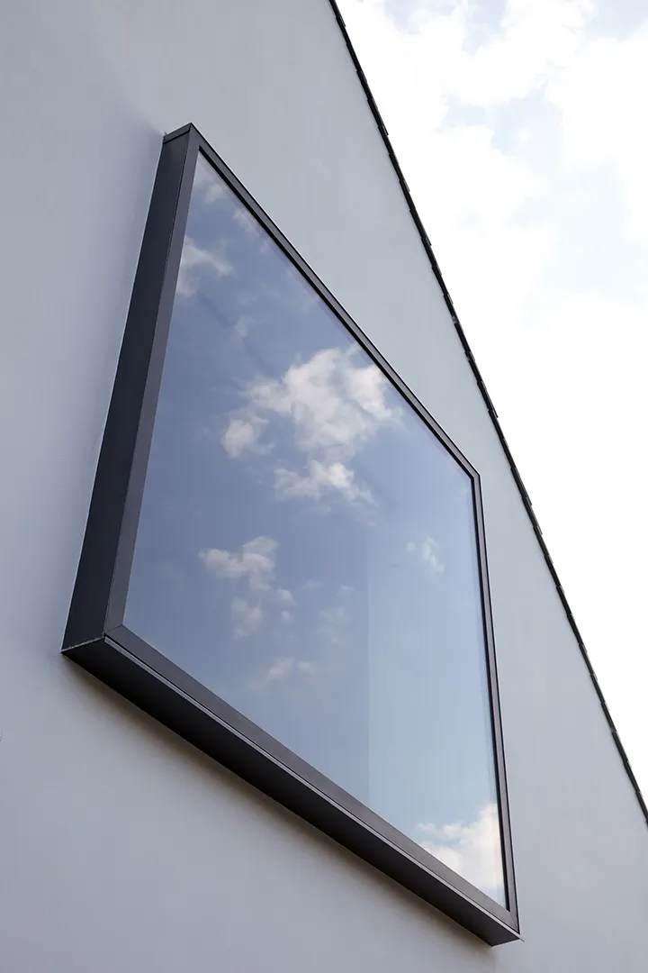 Nahaufnahme eines quadratischen nach außen auskragenden Metallfensters in der weißen Putzfassade aus der Untersicht; im Glas spiegeln sich Wolken, die schräge Trauflinie führt das Auge zur Gebäudeecke.