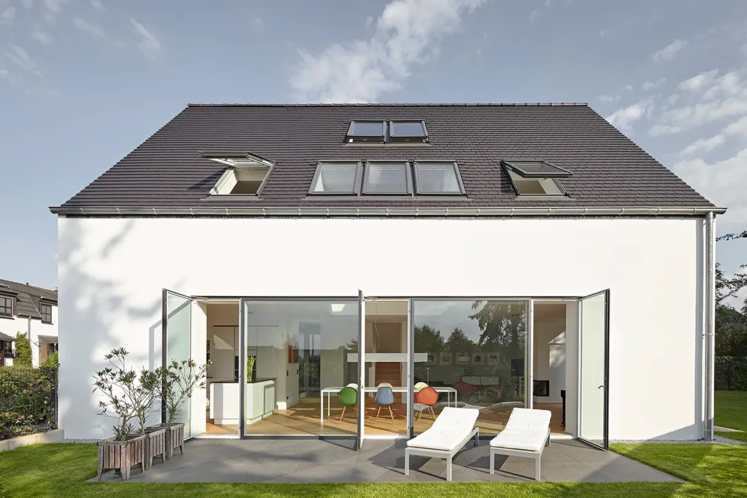 Gartenseitige Außenansicht des traufseitigen Baukörpers; eine raumhohe Faltglasfassade öffnet den Ess- und Wohnbereich vollständig zur Terrasse, das ausladende Satteldach mit mehreren Dachflächenfenstern schließt an die schlichte Putzfassade an.