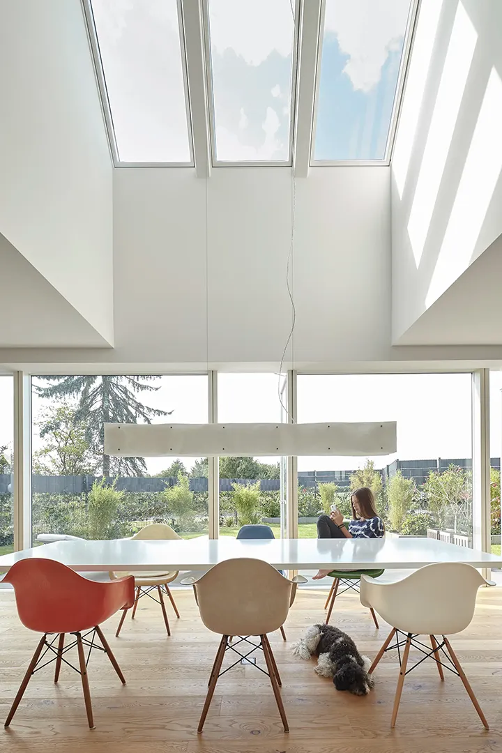 Mehrere Dachflächenfenster fluten den zweigeschossigen Essbereich mit Zenitlicht; die abgeschrägte Dachuntersicht und die umlaufende Rahmenverglasung zum Garten lösen die Raumgrenzen auf. Ein Hund liegt unter dem Esstisch.
