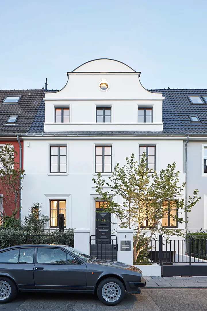 Straßenseitige Ansicht des sanierten Reihenhauses zur blauen Stunde, geschwungener Volutengiebel mit Ochsenauge über symmetrisch gegliederter Lochfassade mit profilierten Fenstergewänden, davor Vorgarteneinfriedung mit Metallzaun.