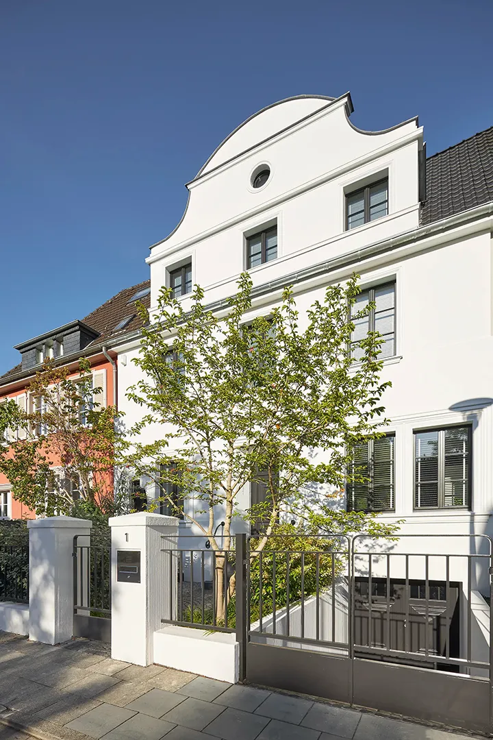 Perspektivische Ansicht der Straßenfassade mit geschwungenem Giebelabschluss und Rundfenster, Fensteröffnungen mit Raffstoren, im Vorbereich Einfriedung mit gemauerten Pfeilern und Zugang zum Souterrain.