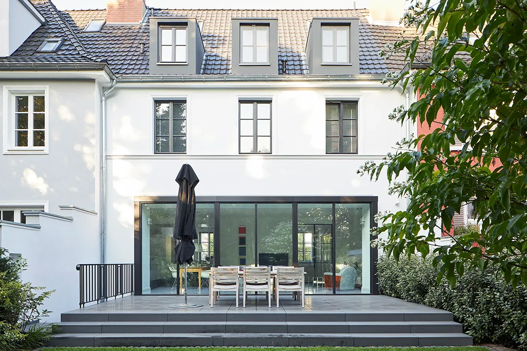 Gartenseitige Ansicht mit vorgesetztem eingeschossigem Anbau in raumhoher Rahmenverglasung, darüber die geputzte Lochfassade mit Schleppgauben im Dachgeschoss, davor Terrasse mit Plattenbelag und Stufenanlage.