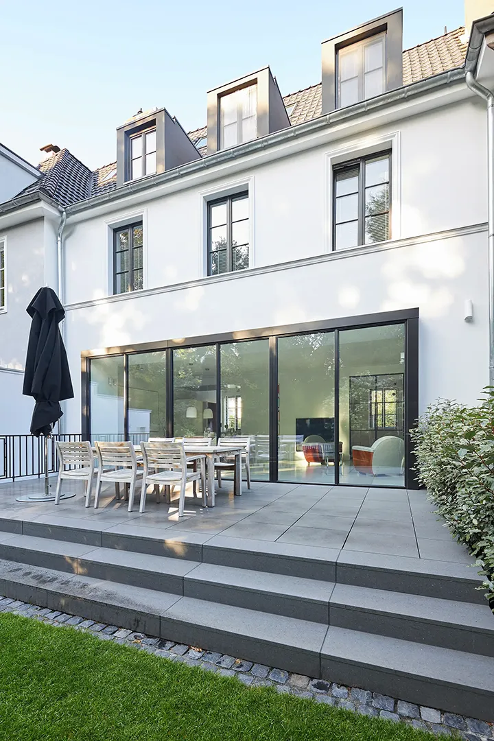 Perspektivische Ansicht des verglasten Anbaus mit dunklen Rahmenprofilen, Terrassenebene mit großformatigem Plattenbelag und umlaufender Stufenanlage in Naturstein, darüber Lochfassade und Dachgauben.