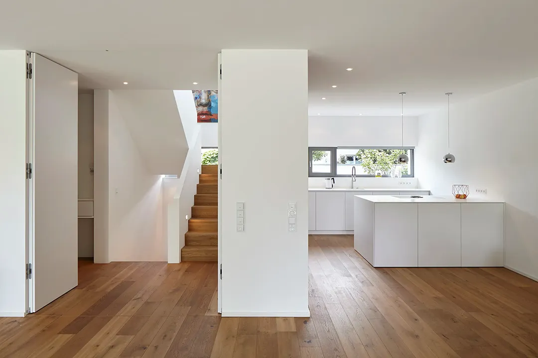 Eine weiße Wandscheibe trennt den offenen Wohnbereich vom Treppenraum; die Holztreppe mit Oberlicht führt ins Obergeschoss, während der angrenzende Kochbereich mit weißen Fronten und Querformat-Fenster sichtbar bleibt.