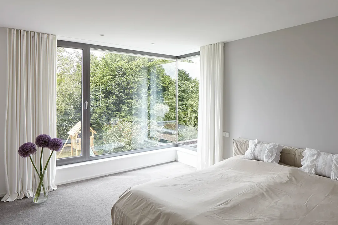 Eckfenster mit fast bodentiefer Rahmenverglasung öffnet zwei Wandflächen des Schlafzimmers zum Grün; die stützenfreie Eckkonstruktion lässt das Fenster nahtlos umlaufen.