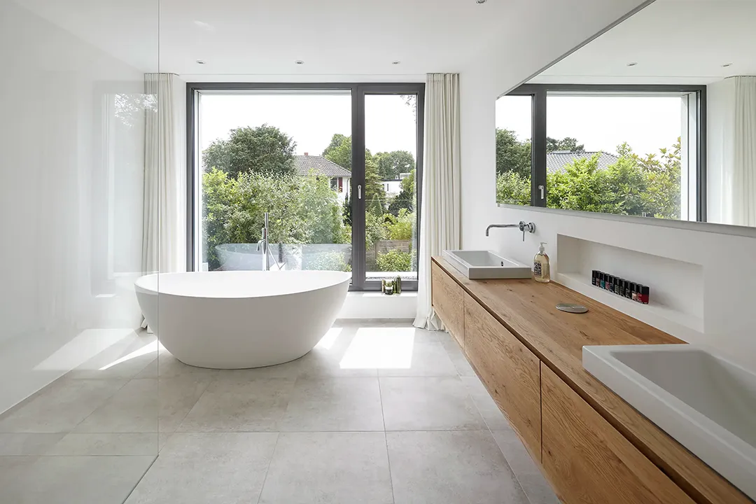 Freistehende Badewanne aus weißem Mineralwerkstoff vor raumhoher Rahmenverglasung mit Gartenblick; ein wandhängendes Holzwaschtischboard mit aufgesetzten Becken und einem langen Spiegel gliedert die gegenüberliegende Wandseite.