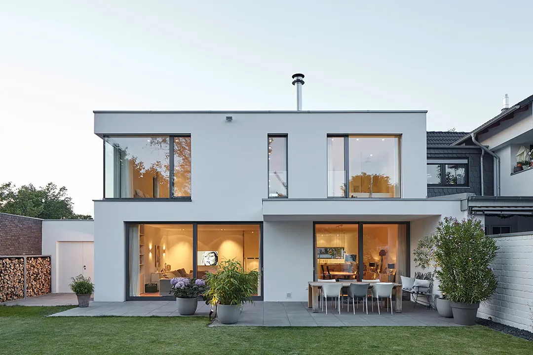 Gartenseitige Außenansicht im Abendlicht: Der zweigeschossige Flachdachbaukörper öffnet sich über großformatige Rahmenfenster und bodentiefe Schiebeverglasung zum Garten; warmes Innenraumlicht zeichnet die Raumgliederung beider Geschosse ab.