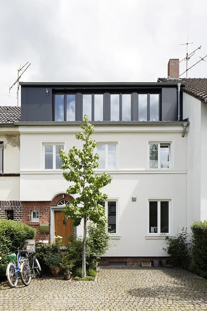 Straßenseitige Außenansicht eines umgebauten Reihenhauses mit weißer Putzfassade; ein durchgehender Dachgeschossaufbau mit umlaufender Rahmenverglasung und dunklem Metallrahmen setzt sich als eigenständiger Körper vom Bestandsbau ab.