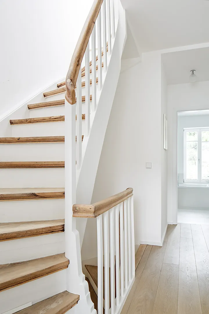 Bestandstreppe mit Holzstufen und weiß gestrichenen Setzstufen und Wangen; der natürlich belassene Holzhandlauf und die Holzbaluster kontrastieren das frisch geweißte Treppenhaus.