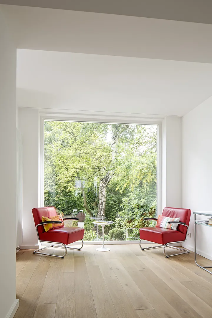 Großformatiges Festglasfenster in tiefer weißer Laibung gibt den Blick in den dicht begrünten Garten frei; die helle Wandfläche und der Holzboden verstärken die Lichtführung im Raum.