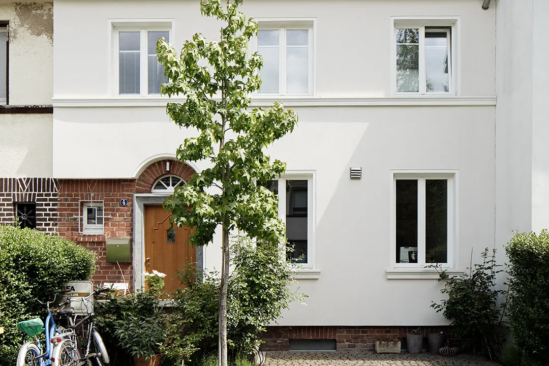 Straßenseitiger Ausschnitt der Bestandsfassade mit weißem Putz über einem Natursteinsockel; ein rundbogiges Eingangsportal aus Naturstein mit Holztür und kleinteilige Sprossenfenster verweisen auf die Bauzeit des Gebäudes.