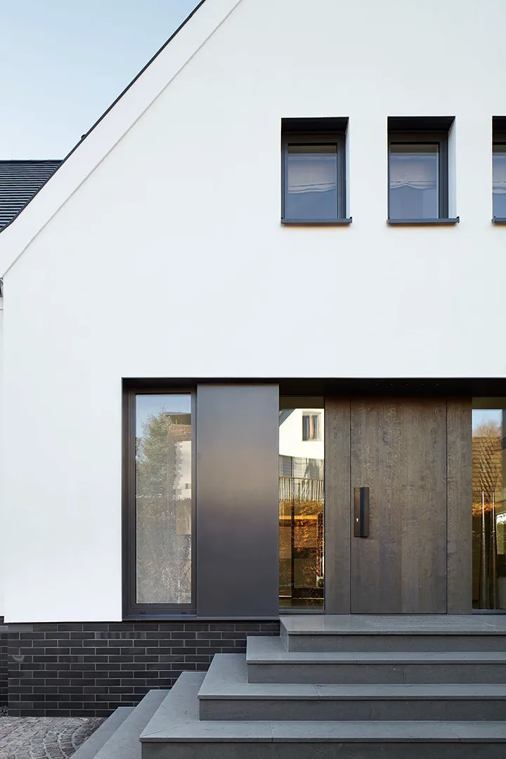 Straßenseitiger Eingangsbereich eines umgebauten Wohnhauses mit weißer Putzfassade und dunklem Natursteinsockel; eine breite Holztür mit seitlicher Rahmenverglasung sitzt flächenbündig in der Fassade, eine Stufenanlage aus dunklem Naturstein führt zum Eingang.