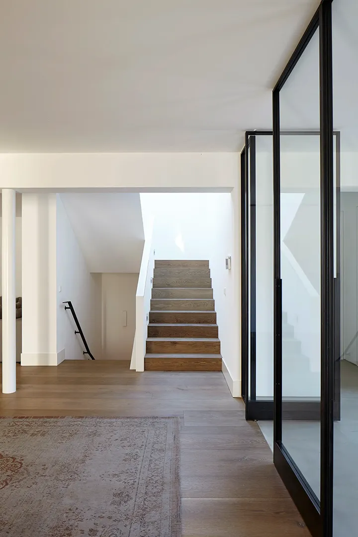 Einläufige Holztreppe in weißem Treppenhaus mit seitlichem Oberlicht; eine raumhohe Stahl-Glas-Tür mit schwarzem Rahmen trennt das Entrée vom angrenzenden Raum.
