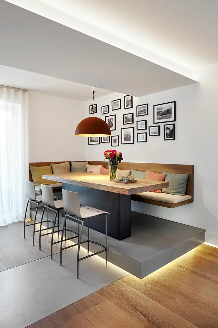 Indirektes Deckenlicht und Sockelbeleuchtung betonen die erhöhte Esszone mit eingebautem Holztisch und umlaufender Wandbank; eine große Hängelampe akzentuiert den Mittelpunkt des Raums.