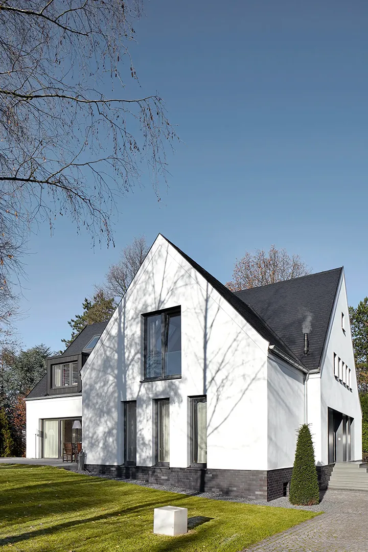 Straßenseitige Außenansicht eines umgebauten Wohnhauses mit steilem Satteldach und weißer Putzfassade über dunklem Natursteinsockel; schmale Hochformatfenster und eine Stufenanlage gliedern den Baukörper im Gegenlicht eines tief stehenden Wintersonnenhimmels.
